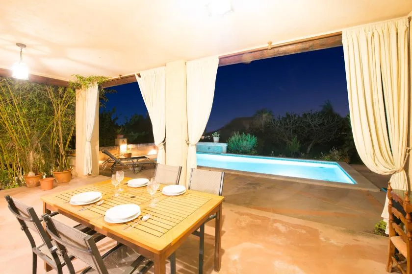 An outdoor dining area beside a lit swimming pool at night.