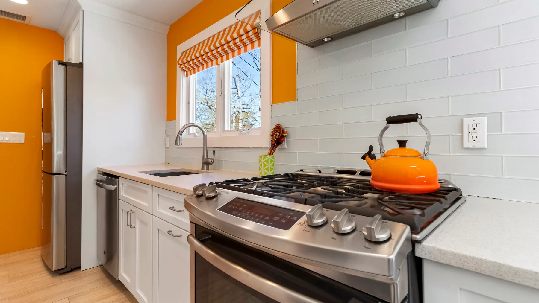 Una cocina luminosa con gabinetes blancos, una estufa de acero inoxidable y una tetera naranja.