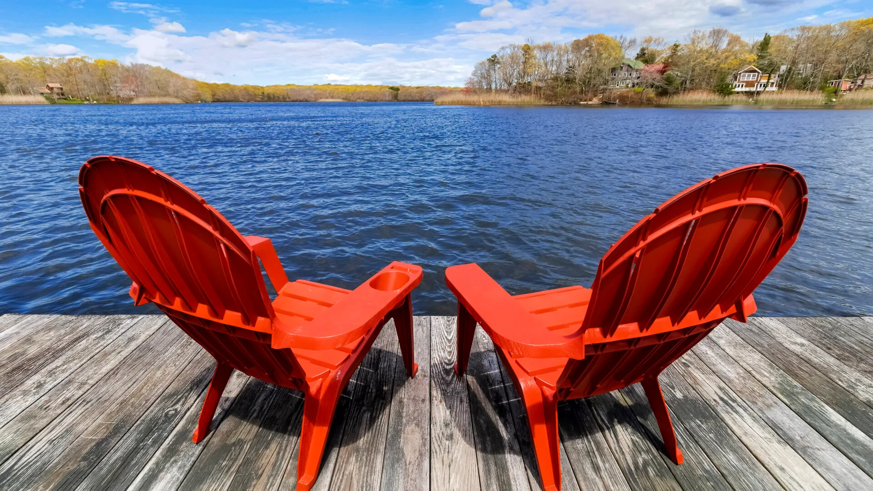 Two red chairs sit on a wooden deck overlooking a lake.