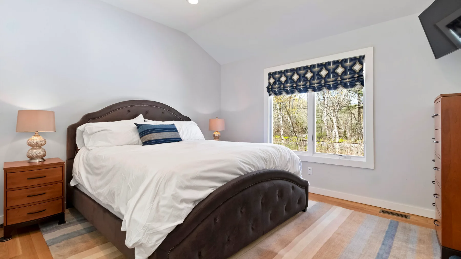 Un dormitorio luminoso con una cama, mesitas de noche y una ventana con vista a los árboles.