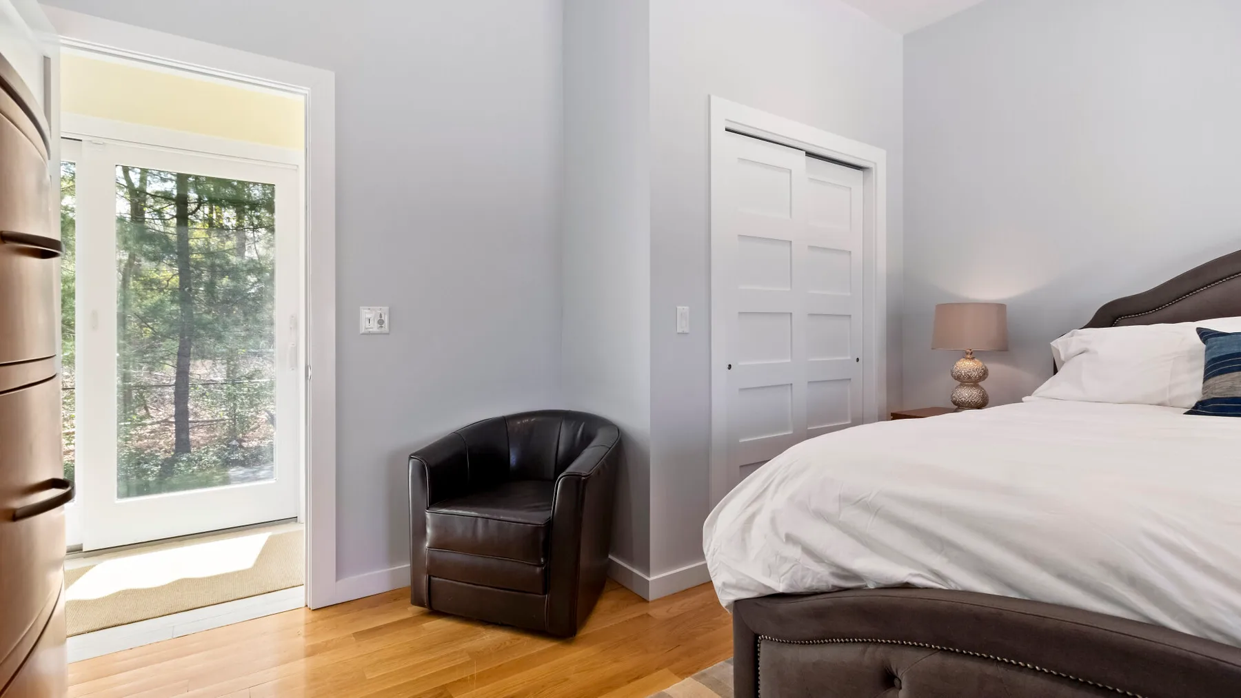 Un dormitorio con una cama, un sillón y una puerta de vidrio con vista a los árboles.
