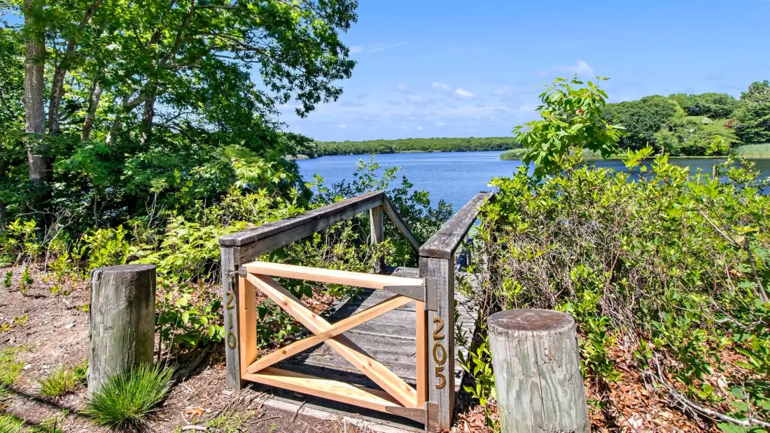 A wooden gate opens onto a path leading toward a lake through leafy greenery.