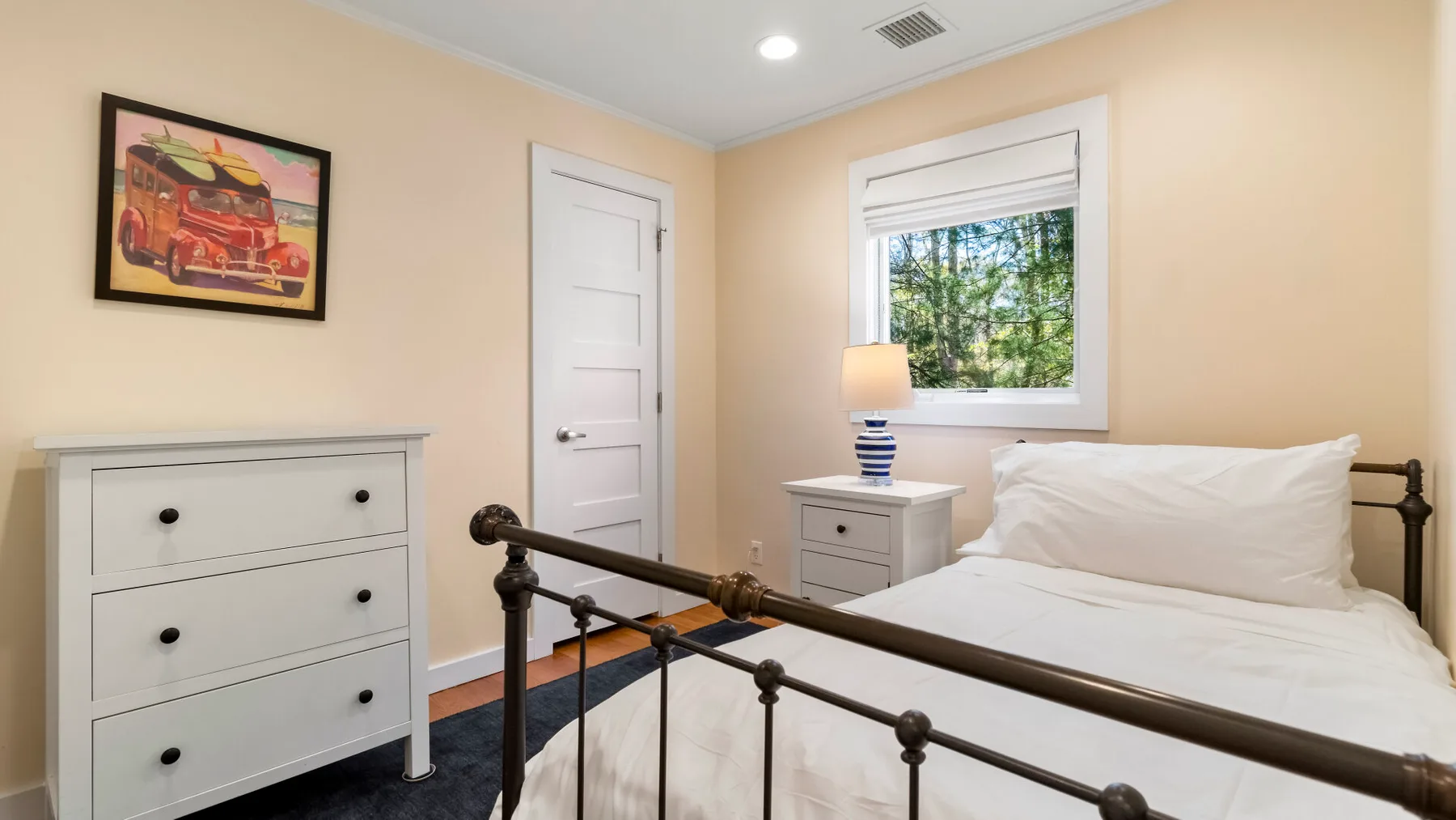 Un dormitorio con una cama blanca, una cómoda y una ventana con vista a los árboles.