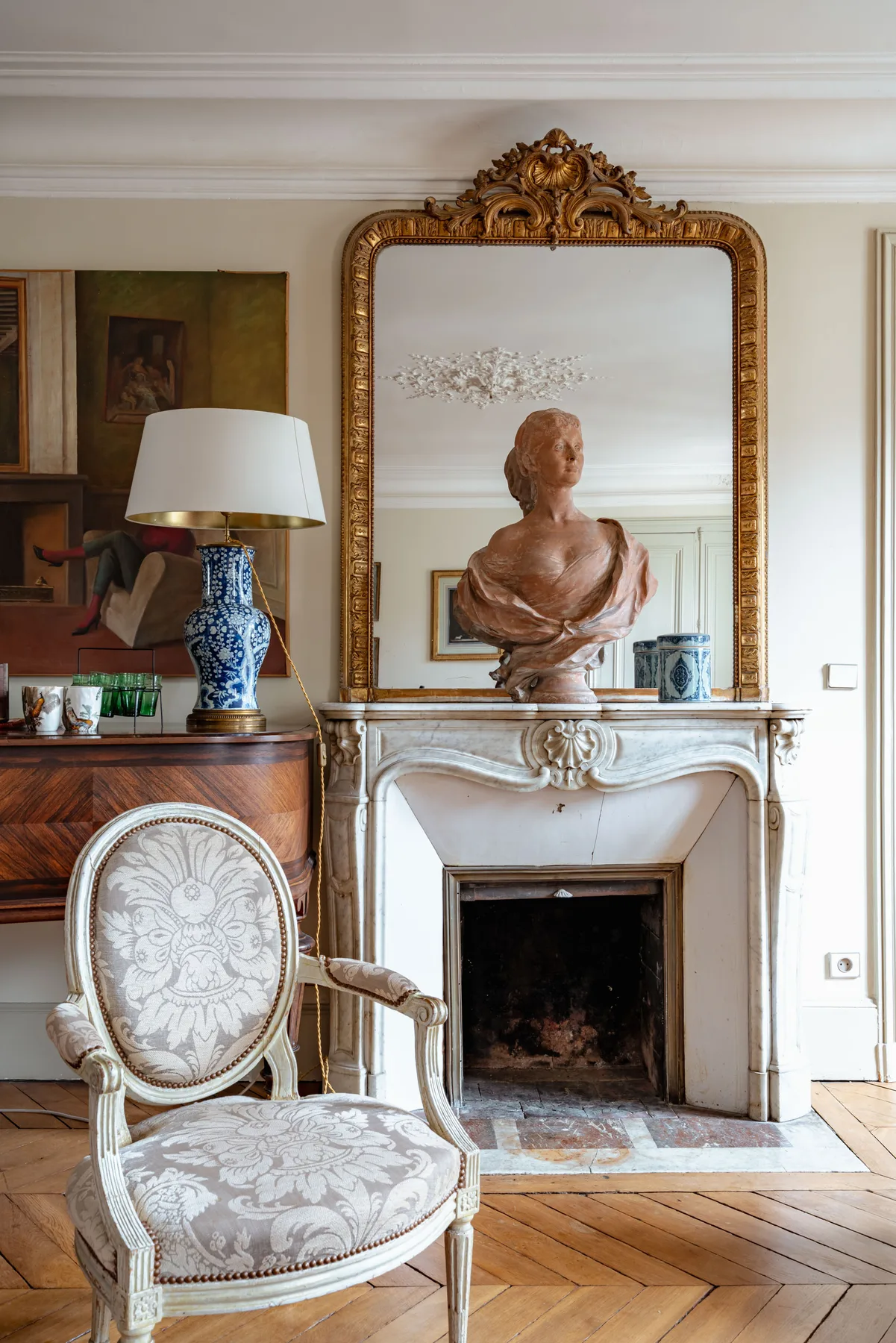 An ornate fireplace and carved mirror stand behind an upholstered chair and bust.