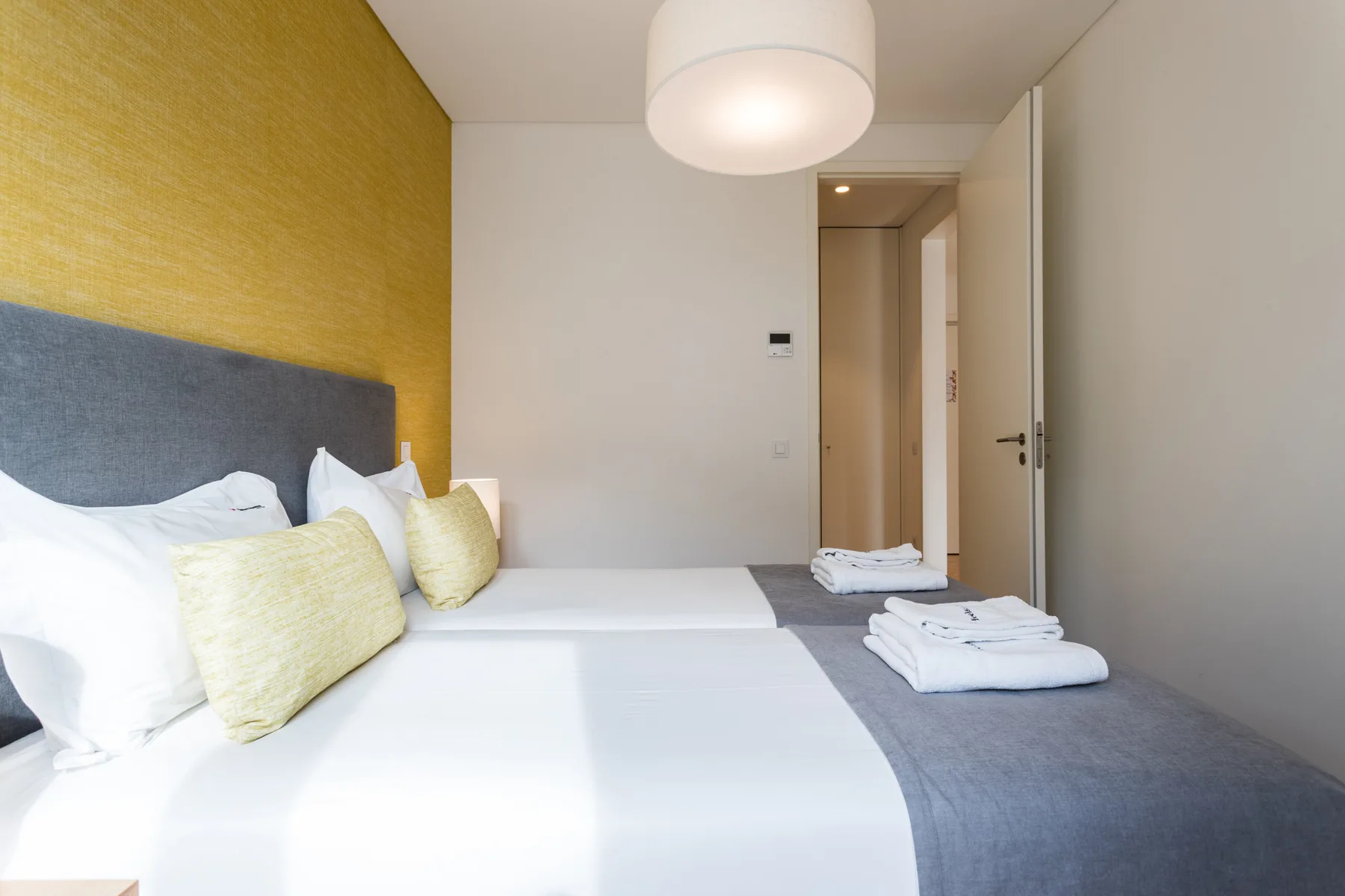 A bedroom with white bedding, pale cushions, and a yellow accent wall.