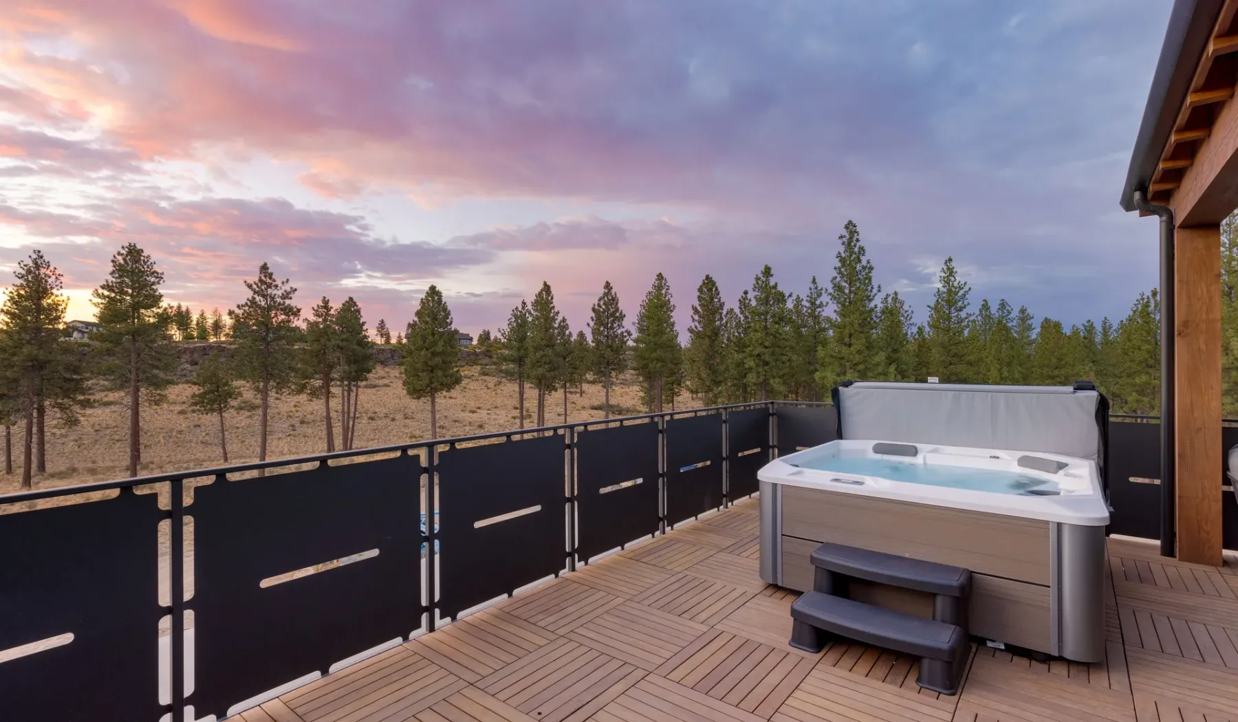 An outdoor hot tub sits on a wooden deck beside a railing, overlooking pine trees.