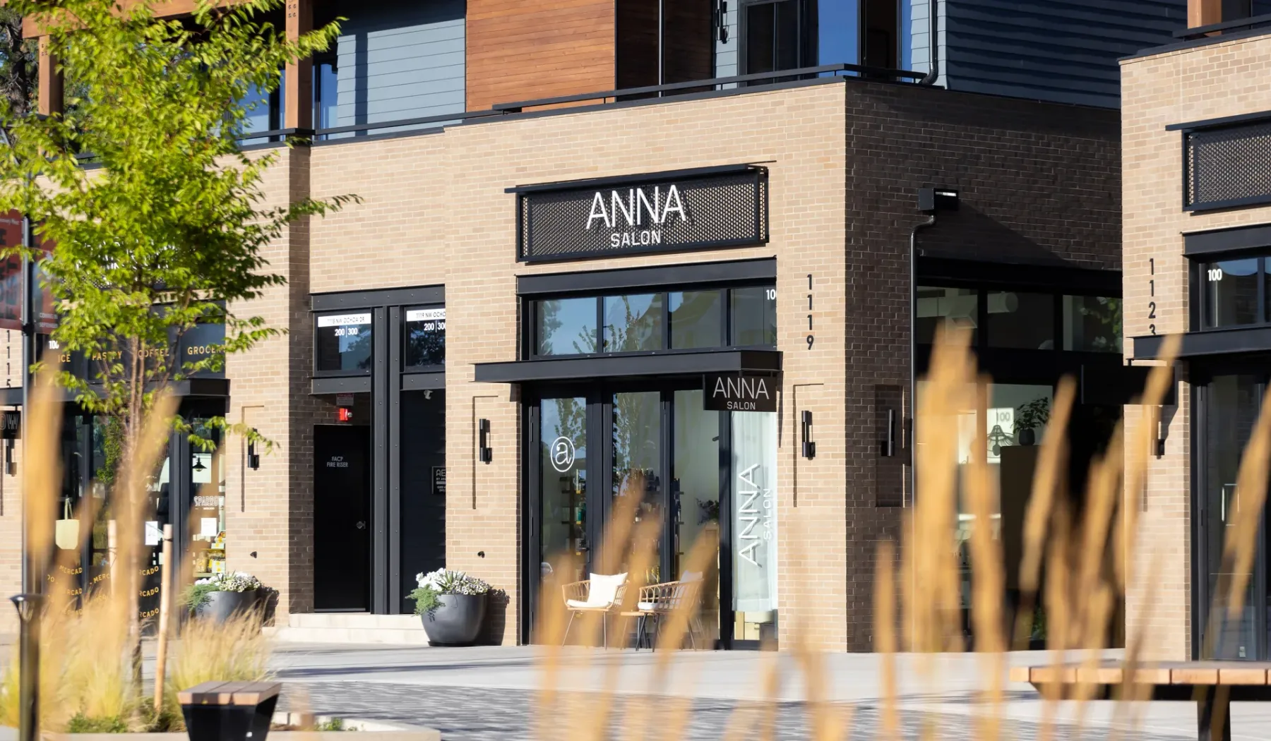 A storefront with large windows and outdoor seating is framed by ornamental grasses.