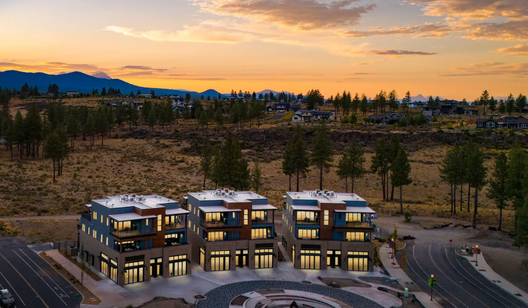 Modern buildings sit beside a broad, tree-dotted landscape beneath a colorful sunset sky.
