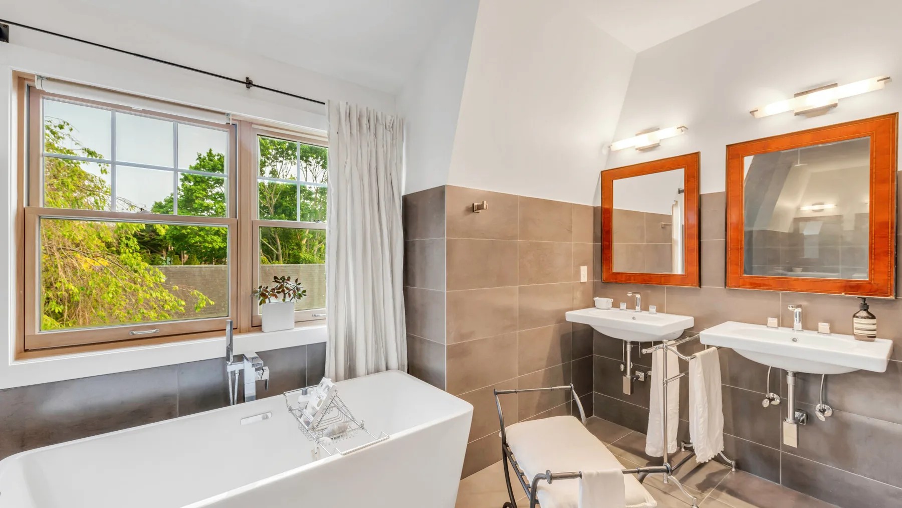 Un baño luminoso con bañera, dos lavabos y grandes ventanas con vistas a la vegetación.
