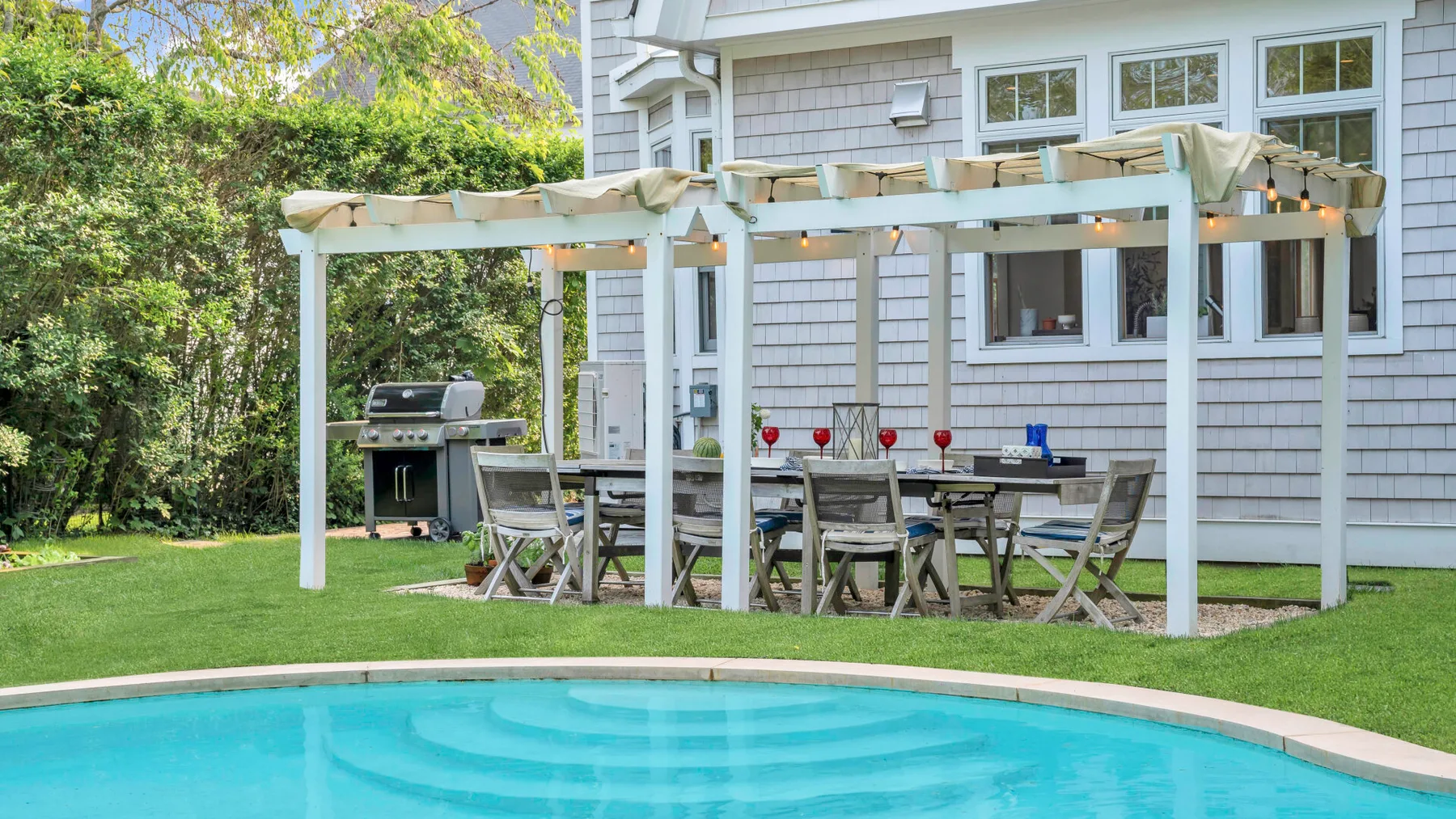 Una piscina al aire libre está junto a un césped y una pérgola con asientos para comer.