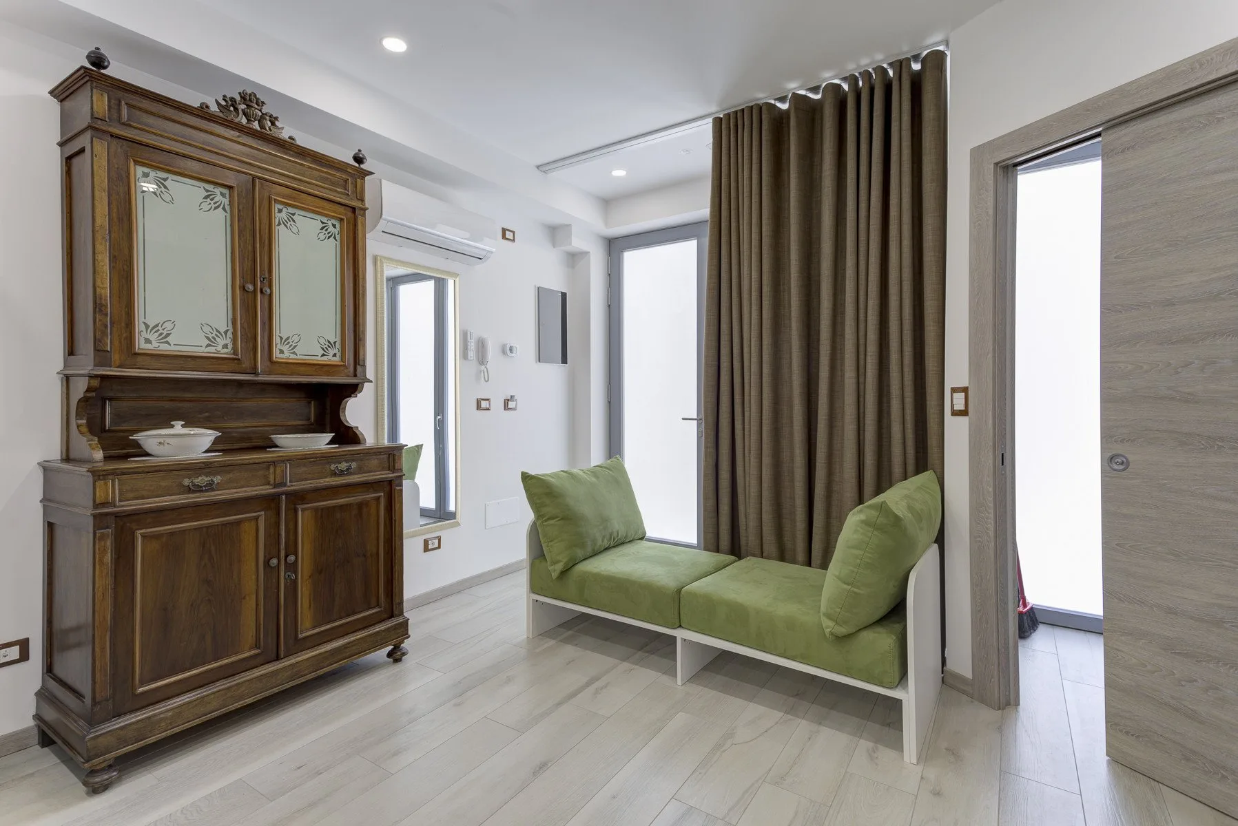 Una zona de estar luminosa con asientos verdes y un armario alto de madera.