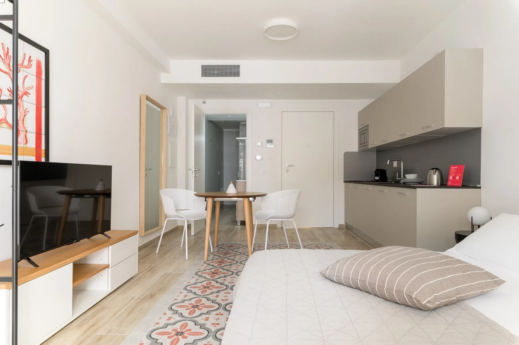 A bright studio room combines a bed, dining table, television, and kitchenette.