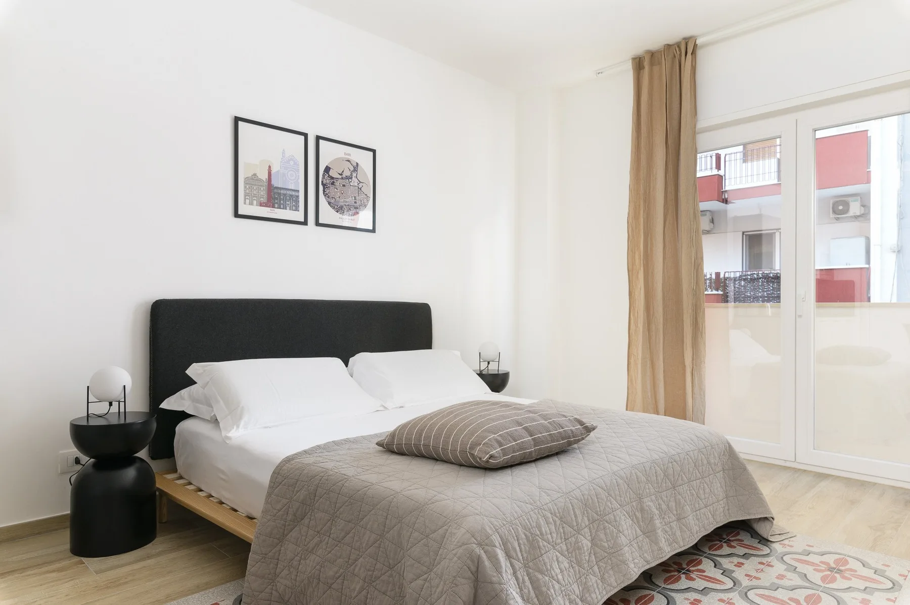 A bright bedroom features a bed with a gray quilt beside a large window.