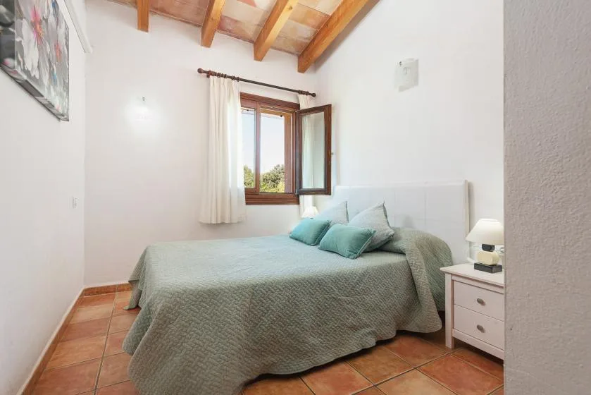 A bedroom with a bed beneath an open window and exposed wooden ceiling beams.