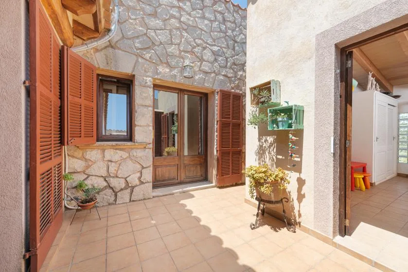 A sunlit stone patio with wooden shutters, a door, and potted plants.