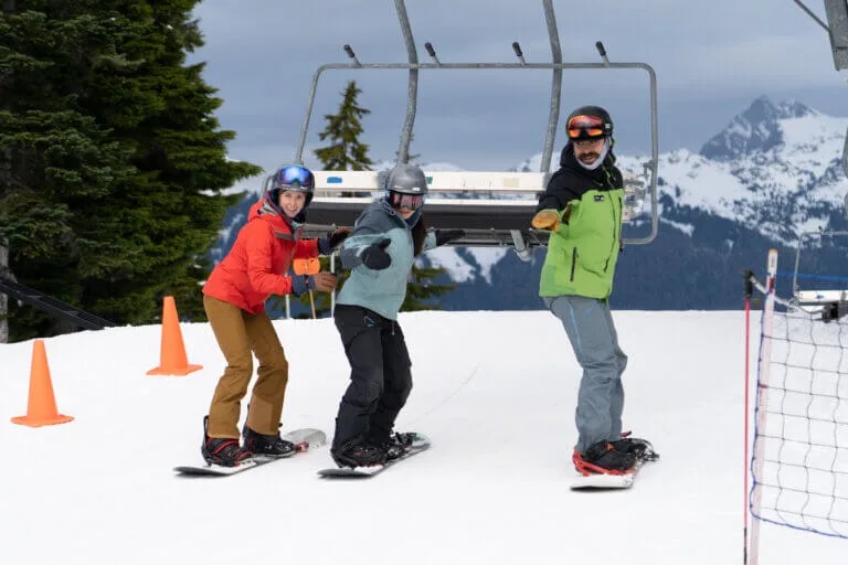 Three snowboarders pause on a snowy slope beneath a chairlift.
