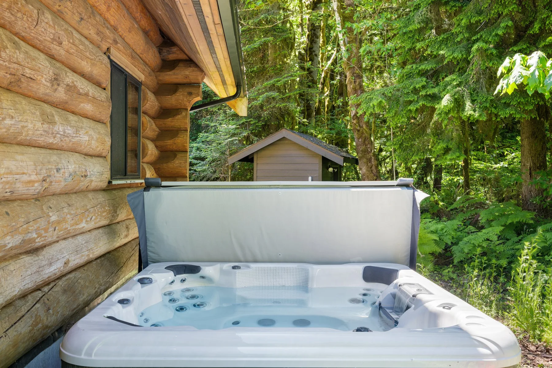 An outdoor hot tub sits beside a log cabin, surrounded by trees and greenery.