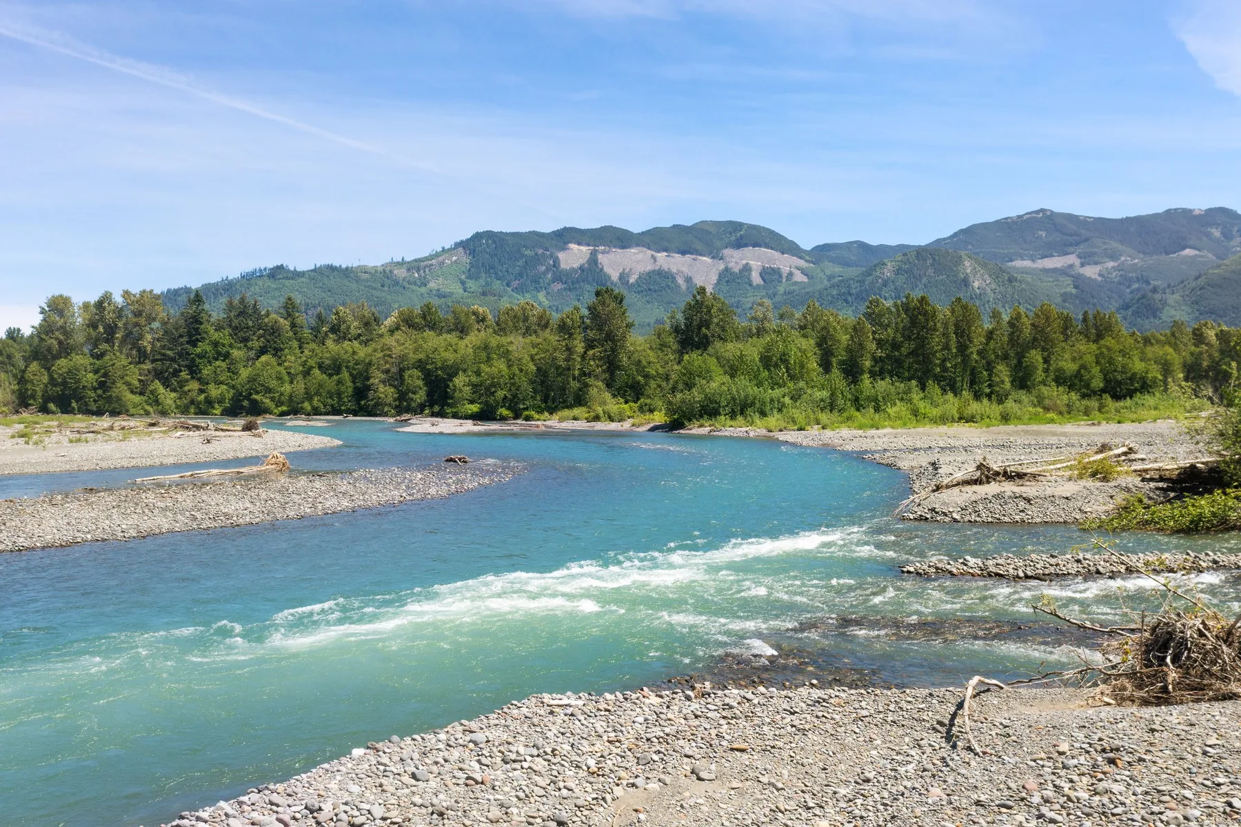 A broad river flows past a gravel bank, with forested hills in the distance.
