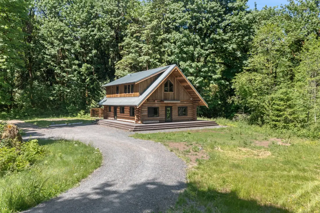 A log cabin stands beside a curving gravel drive, surrounded by trees and grass.