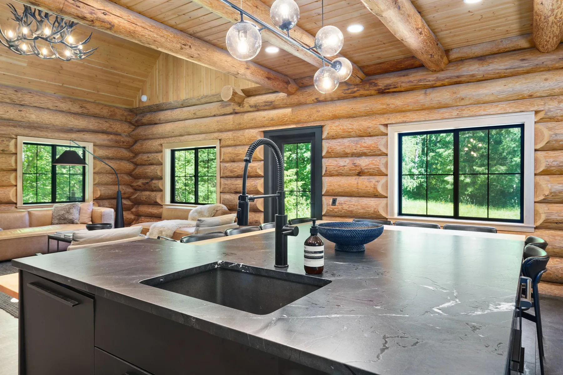 A log cabin kitchen features a large island with a sink and windows overlooking greenery.