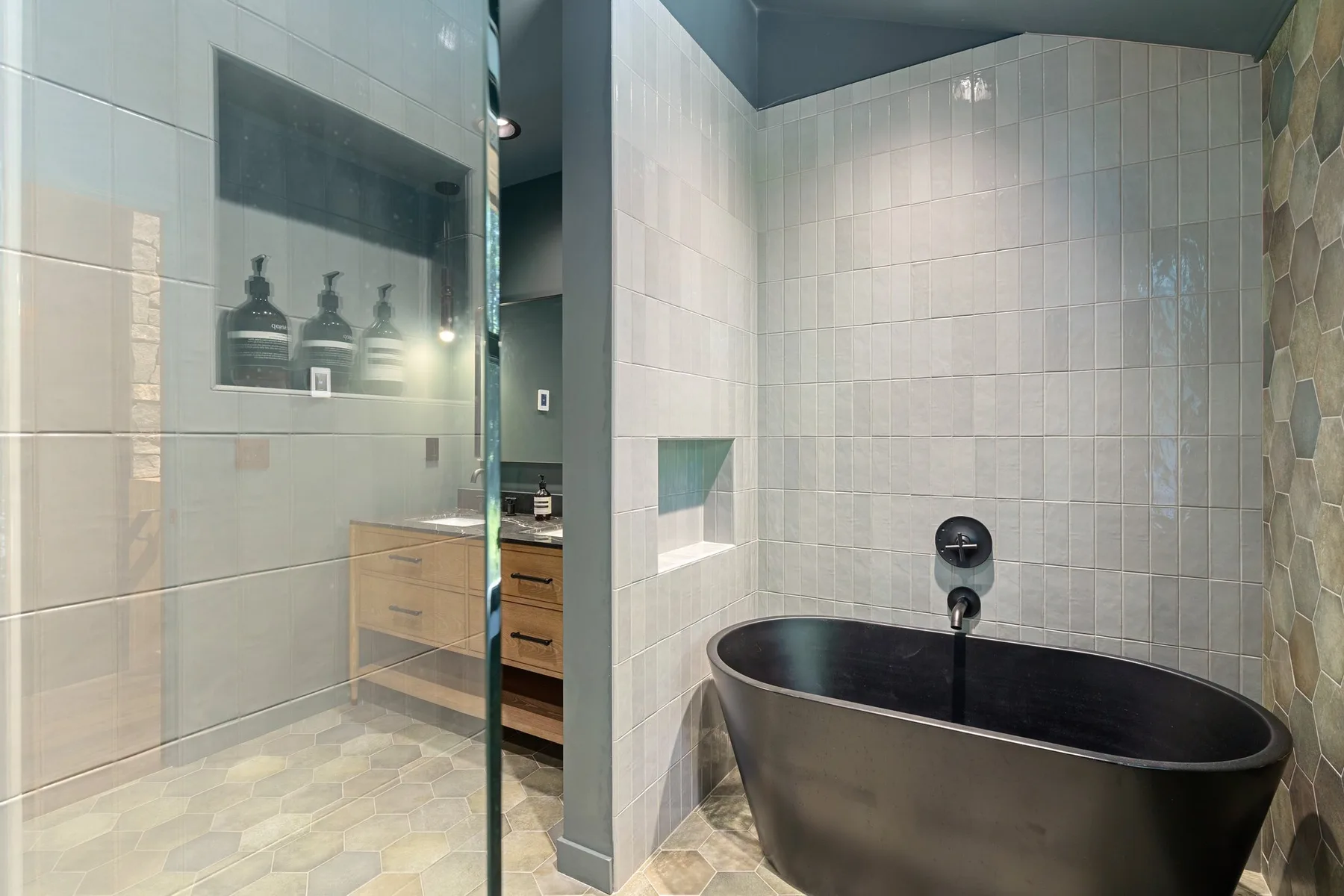 A tiled bathroom with a dark freestanding bathtub and glass shower screen.