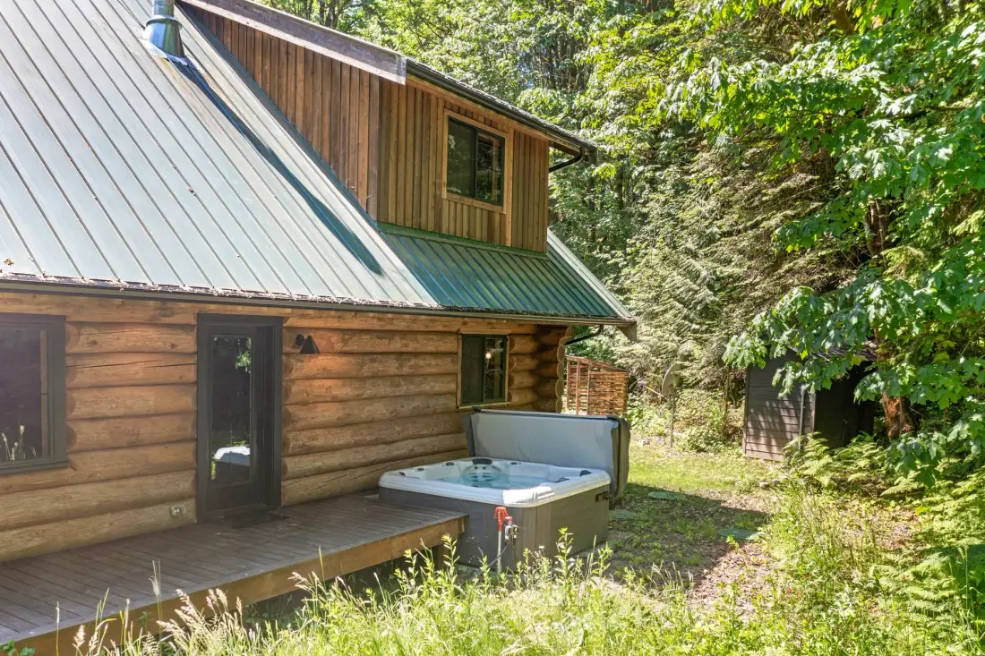 A wood-sided cabin with a metal roof sits among trees, with a hot tub beside the deck.