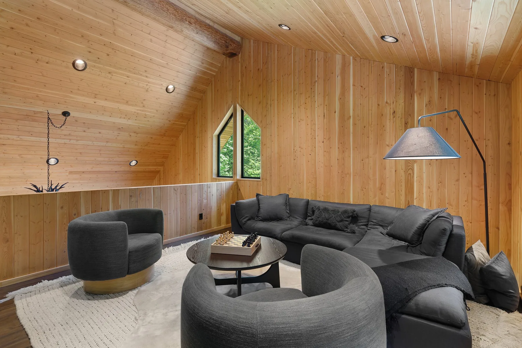 A living room with dark seating and a large floor lamp beneath a sloped wood ceiling.
