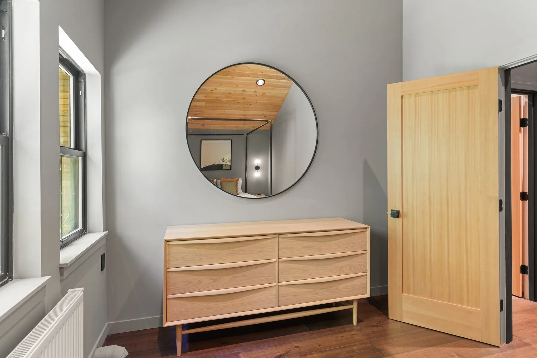 A bedroom with a wooden dresser, round mirror, and an open door.