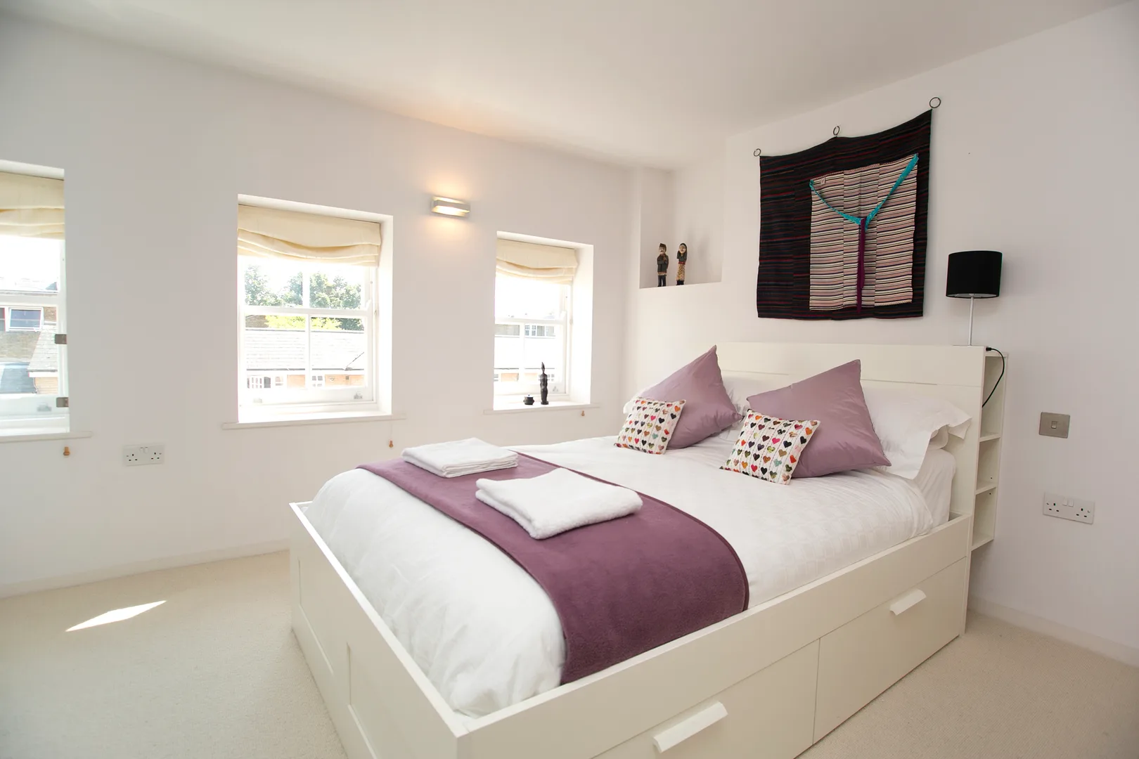 A bedroom with a white bed dressed in white and mauve linens beside bright windows.