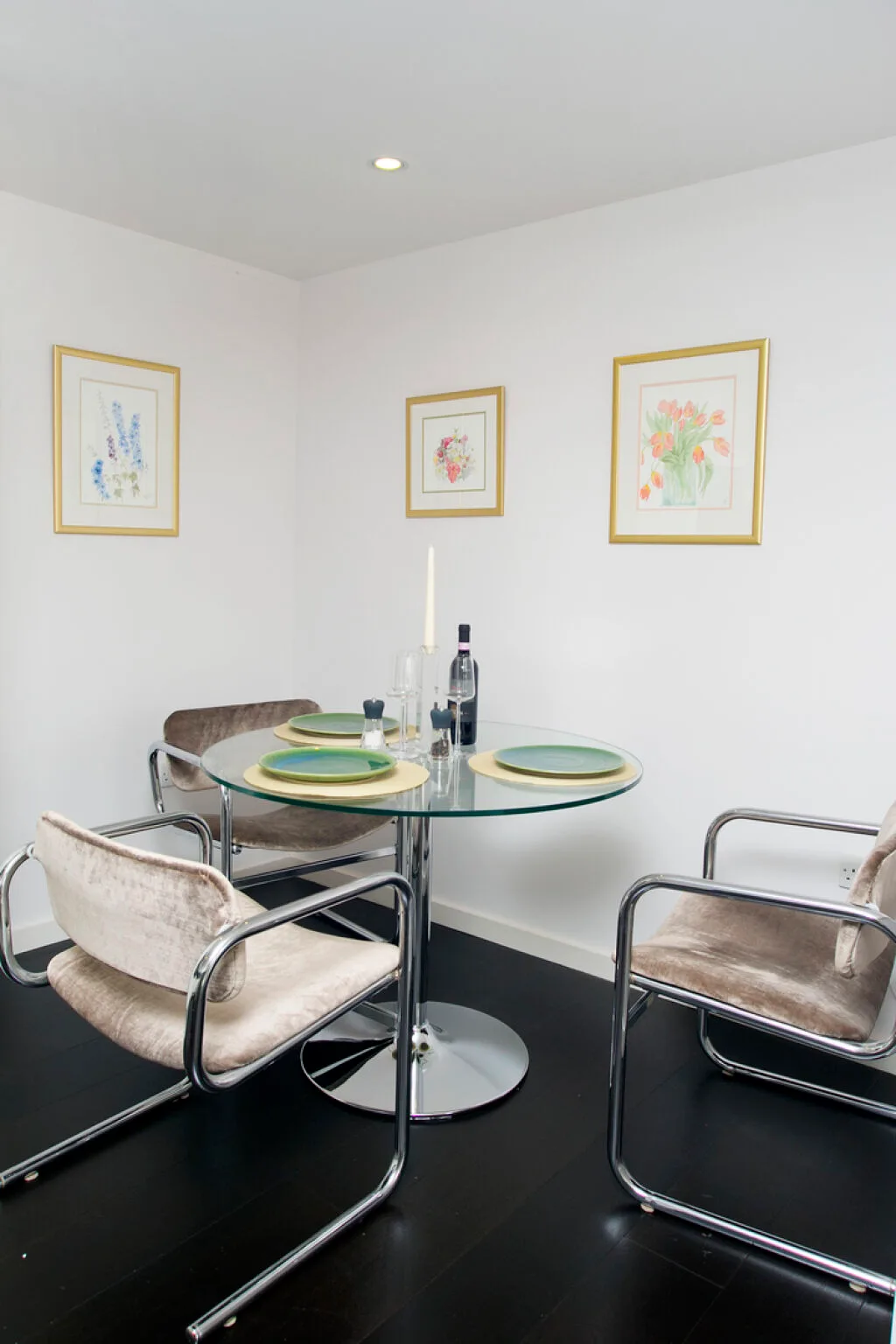 A small dining area with a round glass table, metal seating, and framed floral prints.