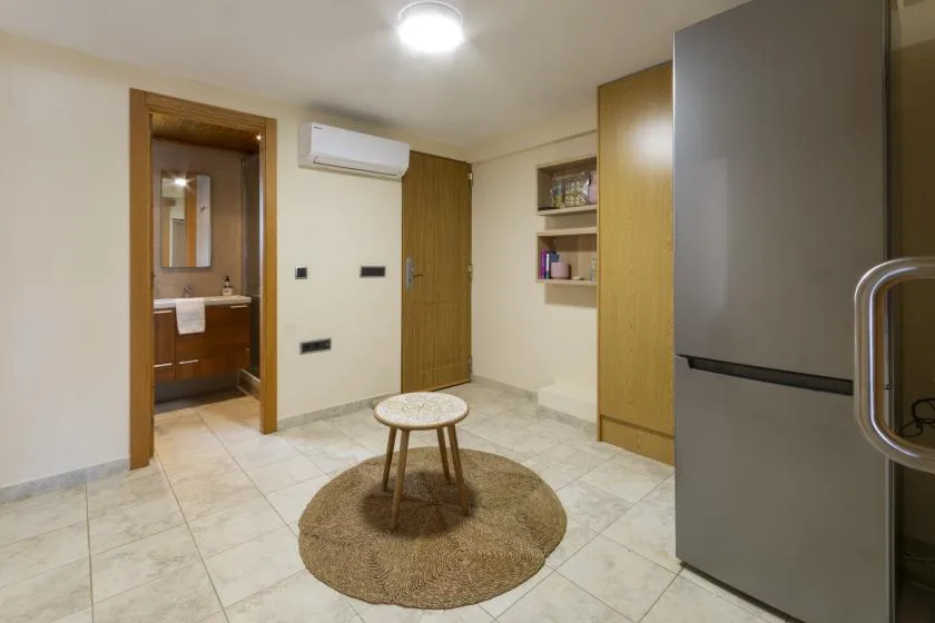 Una habitación con suelo de baldosas, una alfombra redonda y una mesita; se ve un baño al fondo.