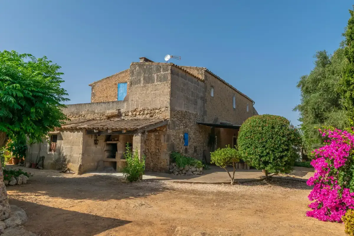 Una casa de piedra rústica se alza en un jardín soleado con arbustos floridos.