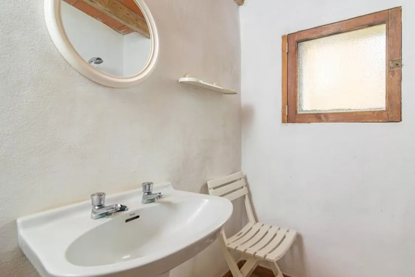 Un lavabo de baño está bajo un espejo redondo, junto a una ventana pequeña y un asiento plegable.