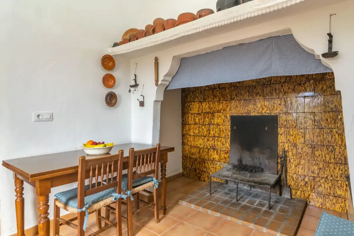 Un comedor junto a una chimenea revestida de azulejos, con mesa y sillas de madera.