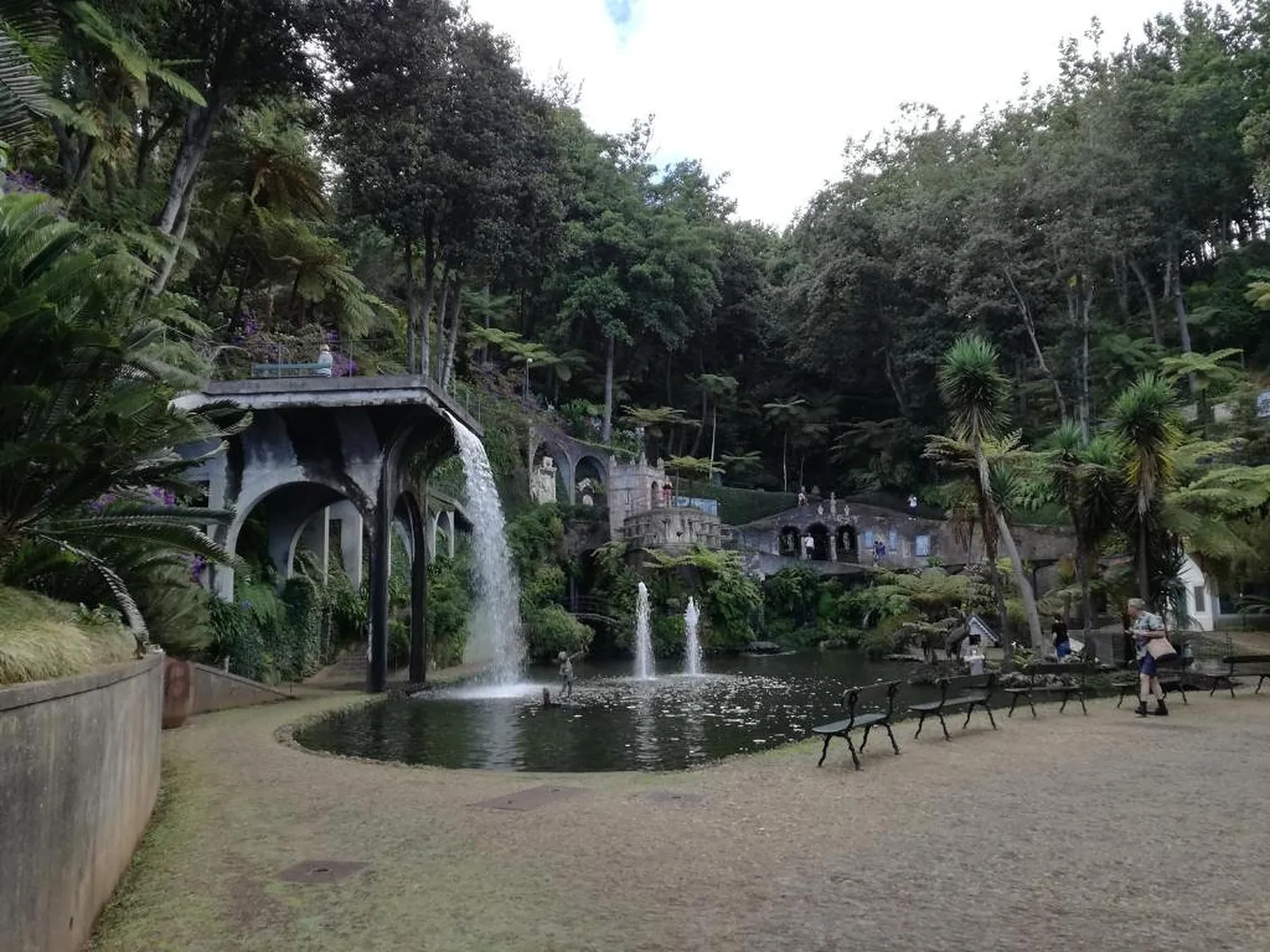Estanque en un parque arbolado con fuentes, una cascada y bancos junto a la orilla.