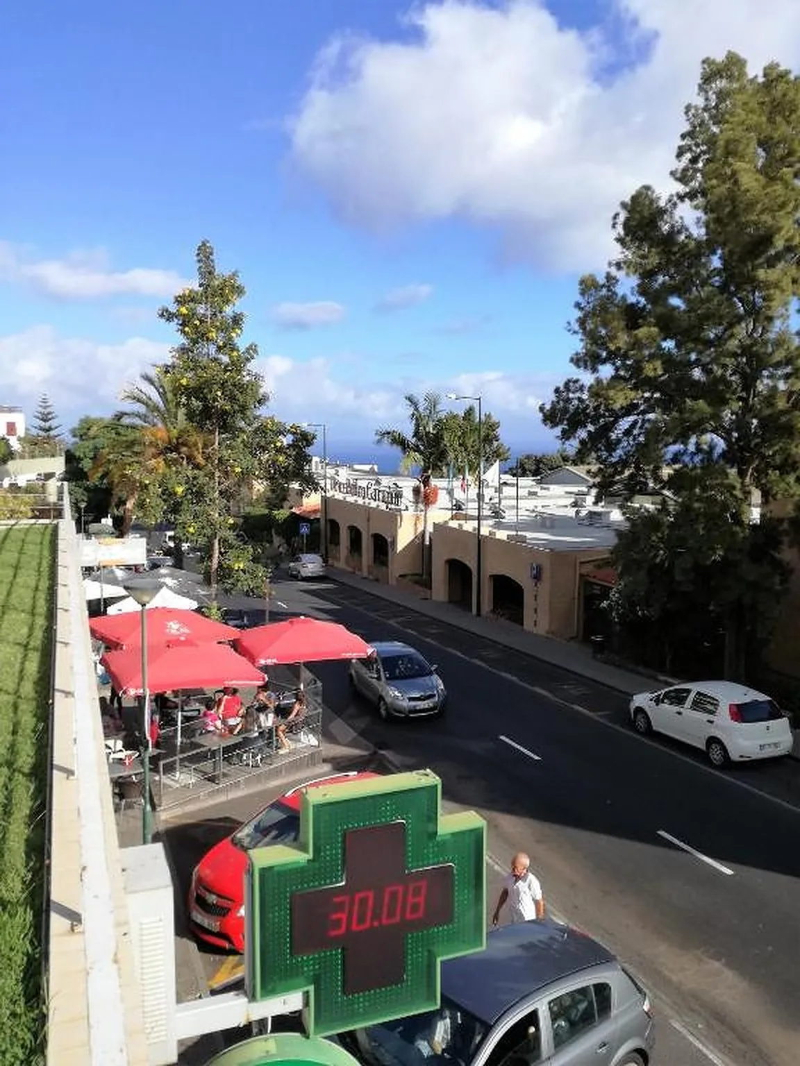 Una calle con autos estacionados, comercios y un letrero de farmacia bajo un cielo azul.
