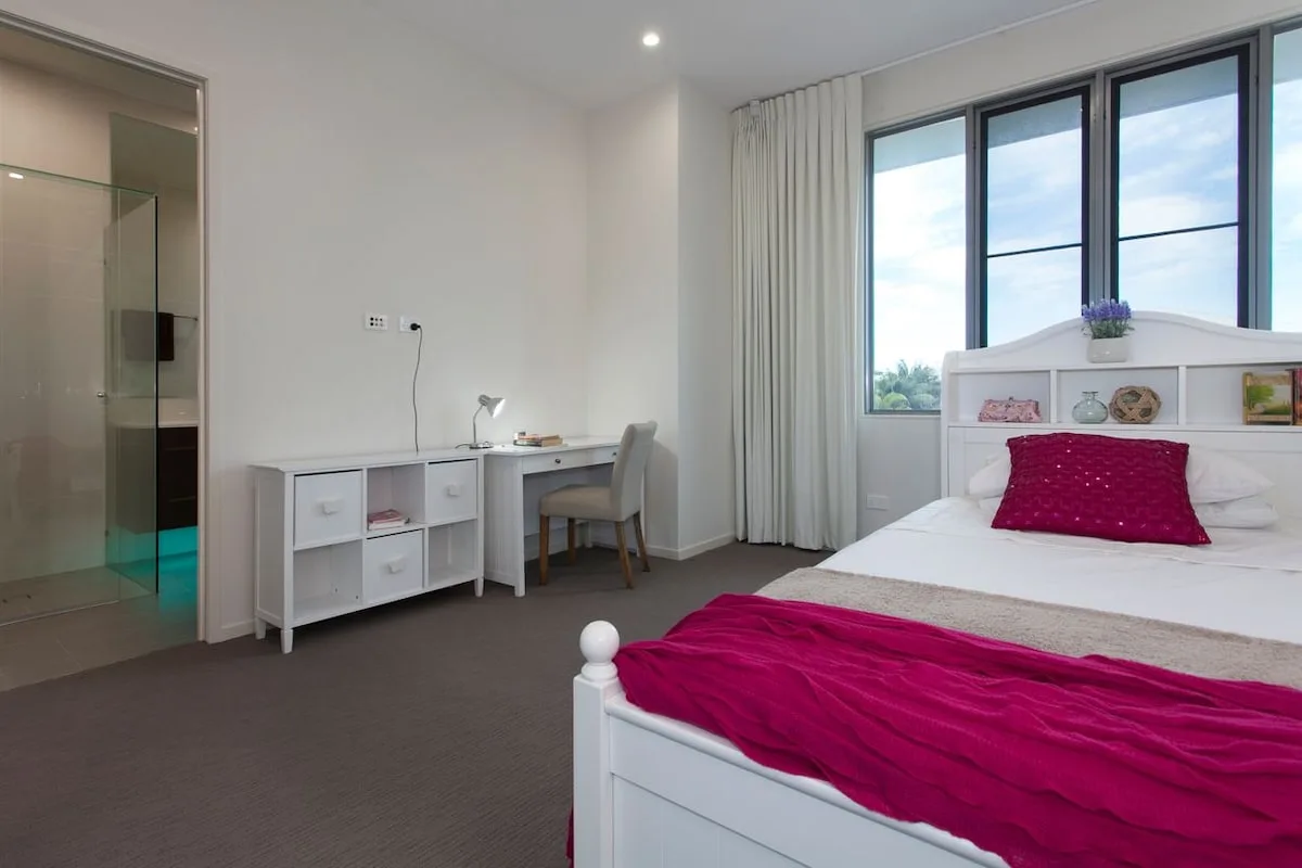 Dormitorio luminoso con una cama blanca y un pequeño escritorio junto a la ventana.