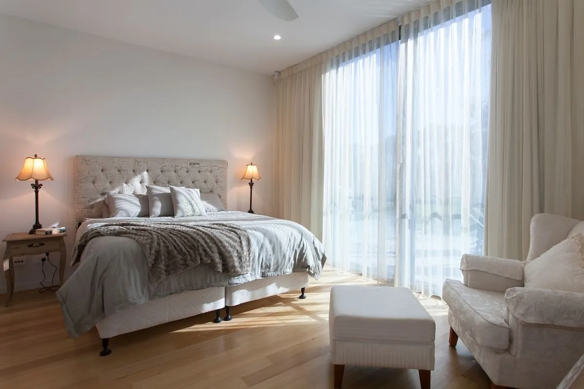 Un dormitorio luminoso con una cama, lámparas de noche y ventanas altas con cortinas.