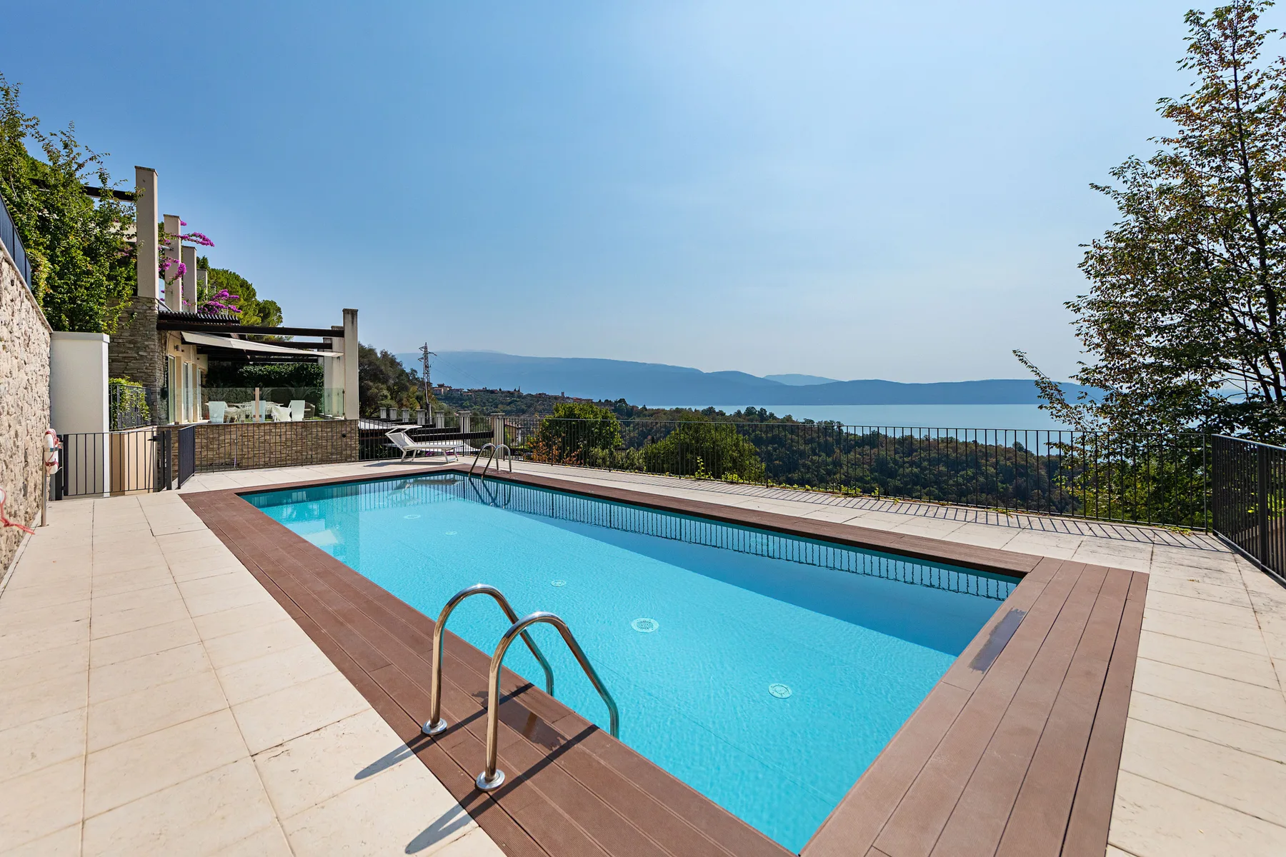 An outdoor swimming pool sits beside a broad deck, with hills and water in the distance.