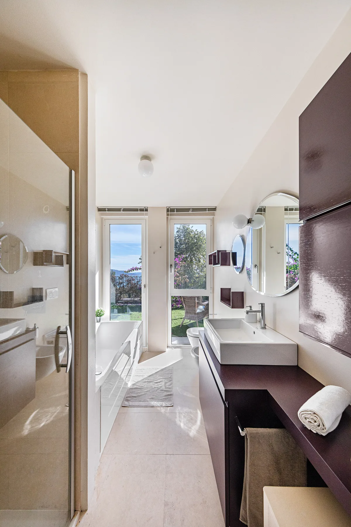 A bright bathroom with a glass shower, bathtub, and vanity beside a garden-facing window.