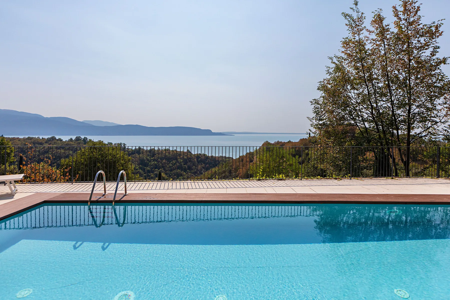 An outdoor swimming pool overlooks a lake and wooded hills.