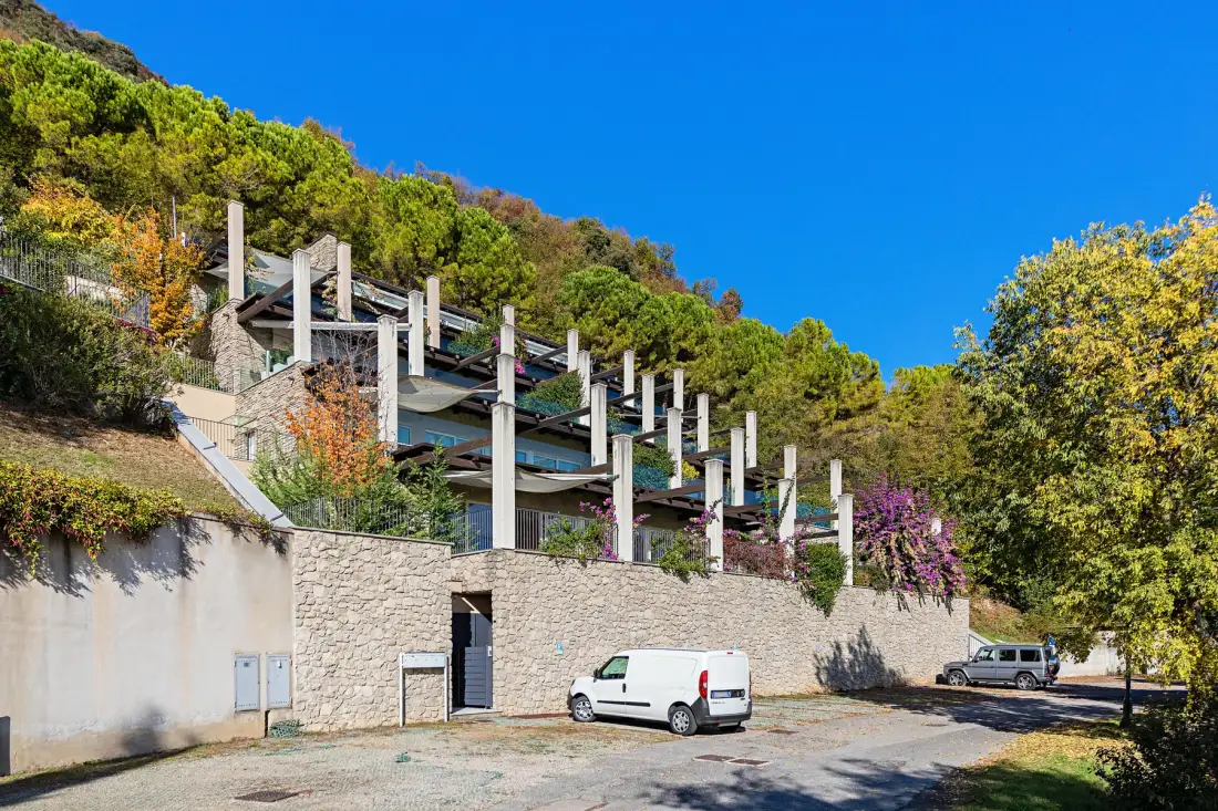 A multi-level building sits against a wooded hillside above a stone retaining wall.