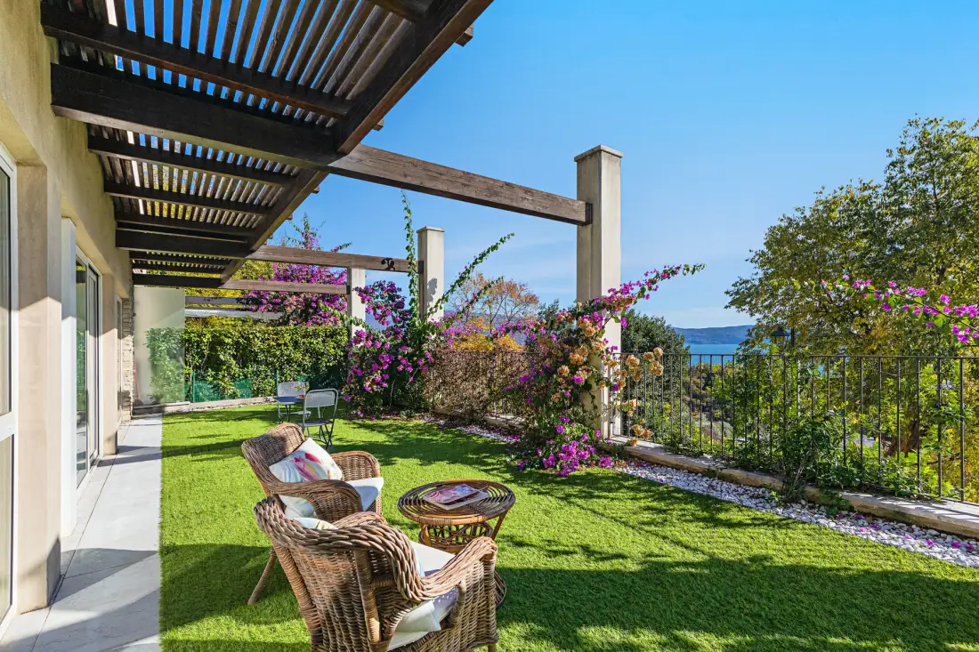 A garden patio with wicker seating, flowering vines, and a view of the water.