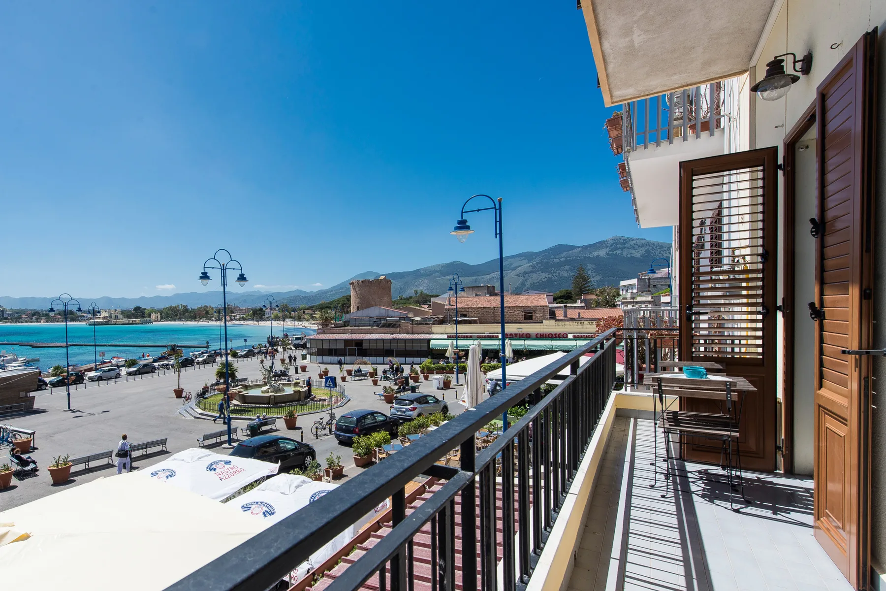 A balcony overlooks a waterfront promenade with the sea and mountains in the distance.