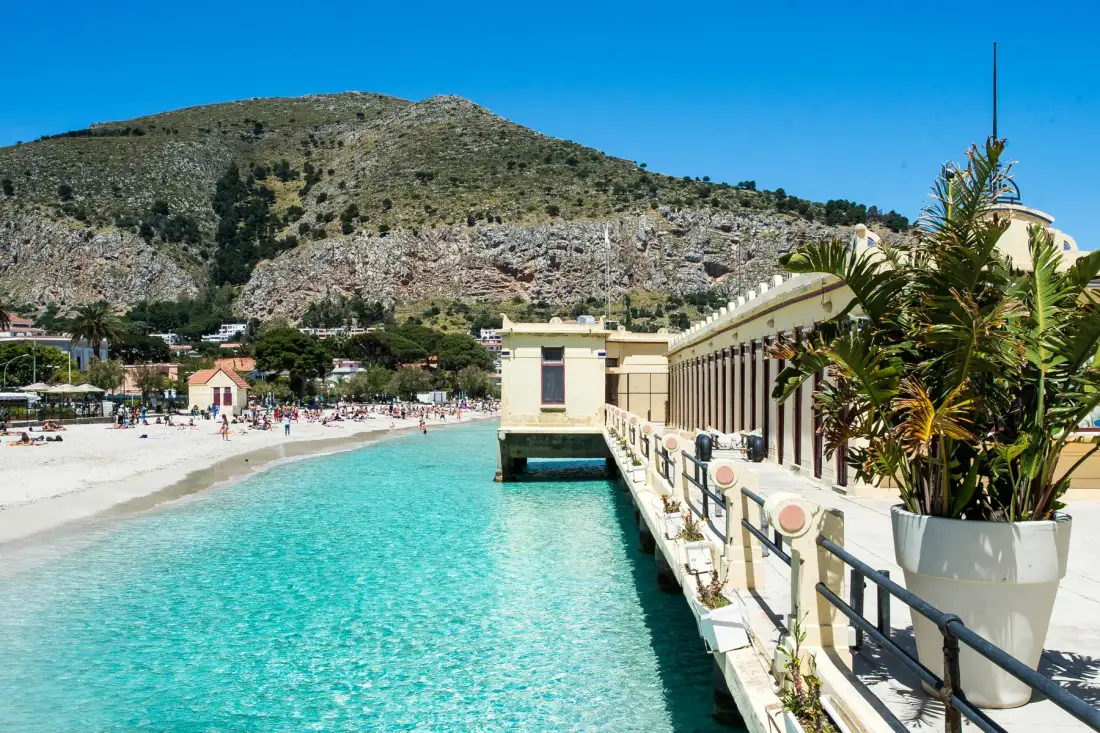 A seaside promenade borders clear turquoise water beside a sandy beach.