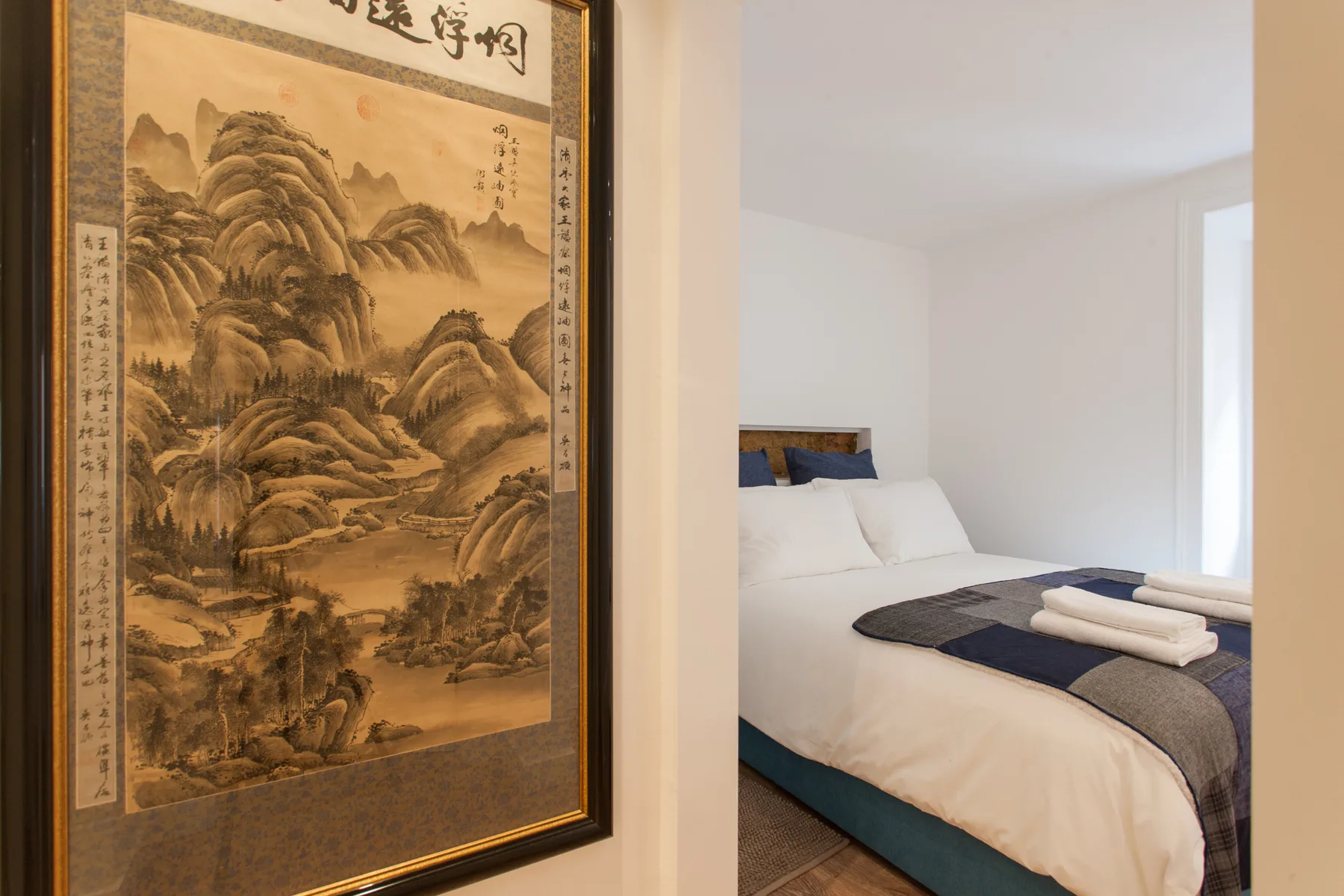 A bedroom with a neatly made bed and a framed landscape painting.