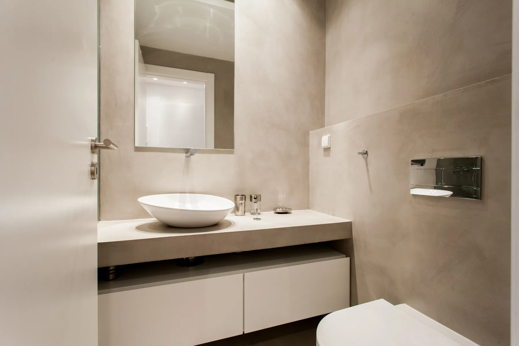 A modern bathroom with a vessel sink, mirror, and wall-mounted flush plate.