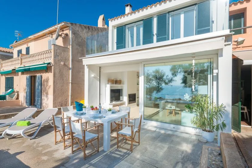A sunlit outdoor dining area sits beside a modern house with large glass windows.