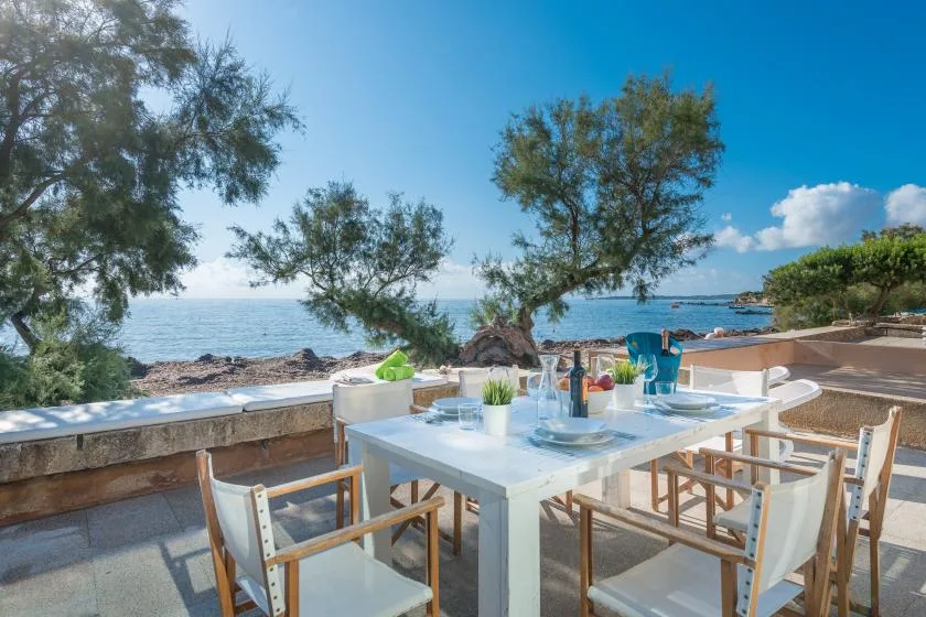 An outdoor dining area overlooks the sea, with a table set beneath leafy trees.