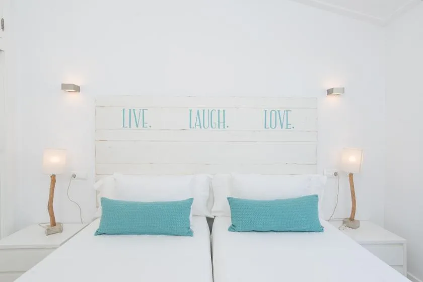 A bright bedroom with two beds and turquoise pillows beneath a white headboard.